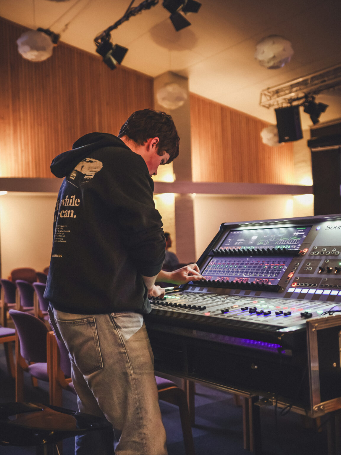 Sam står ved en Soundcraft-mixerpult i en tom sal og gør klar før et arrangement.